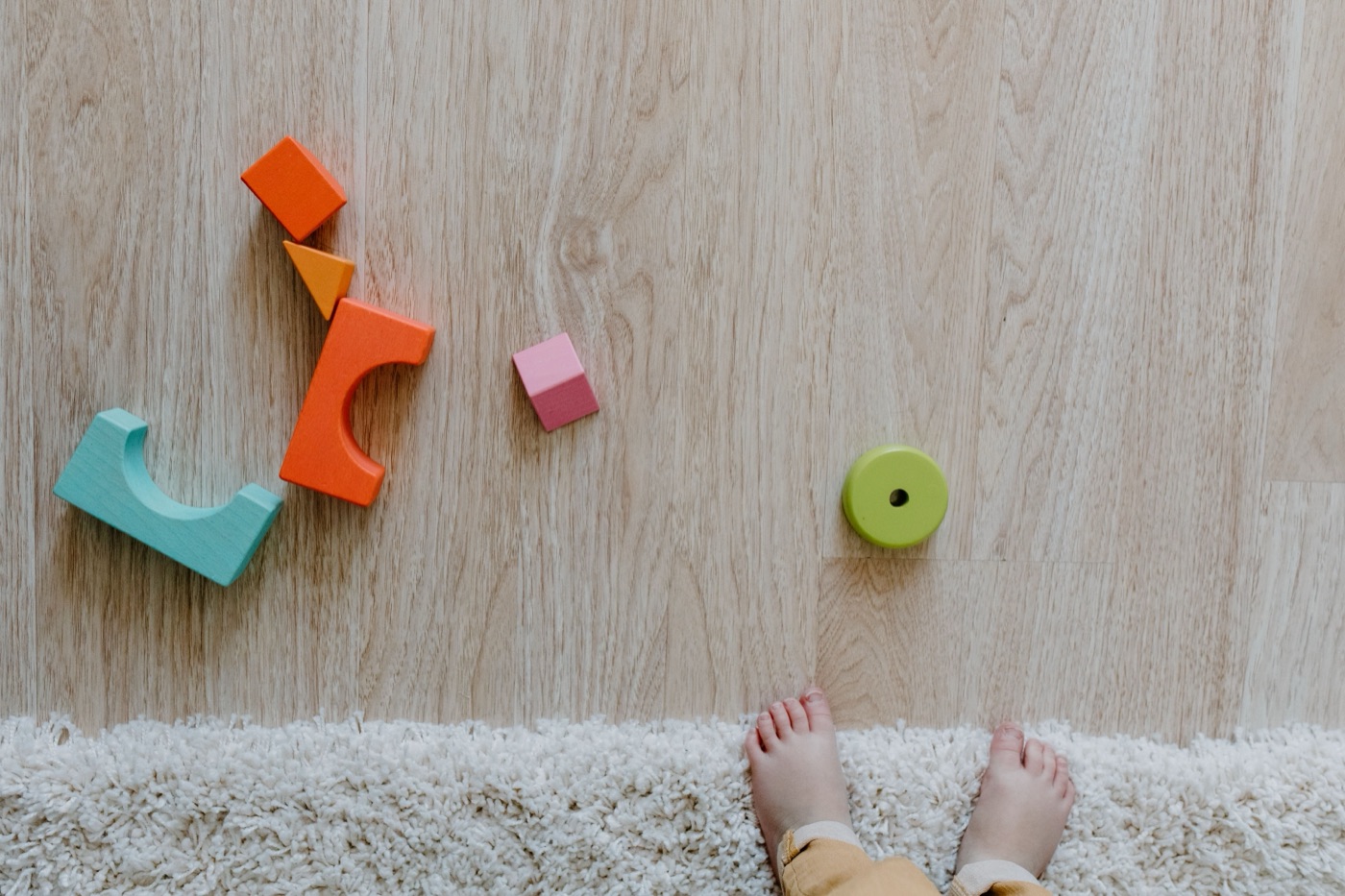 Colorful wooden Montessori blocks on a wood floor beside a toddler's bare feet on a soft rug.