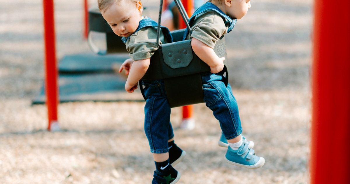 Two babies swinging together on a playground swing.